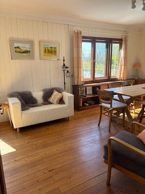 A room with a white sofa, wooden dining table, and chairs, with a view of greenery outside. - Holiday Home 'Kunterbunt' with Private Terrace, Shared Garden and Wi-Fi (Lindau)