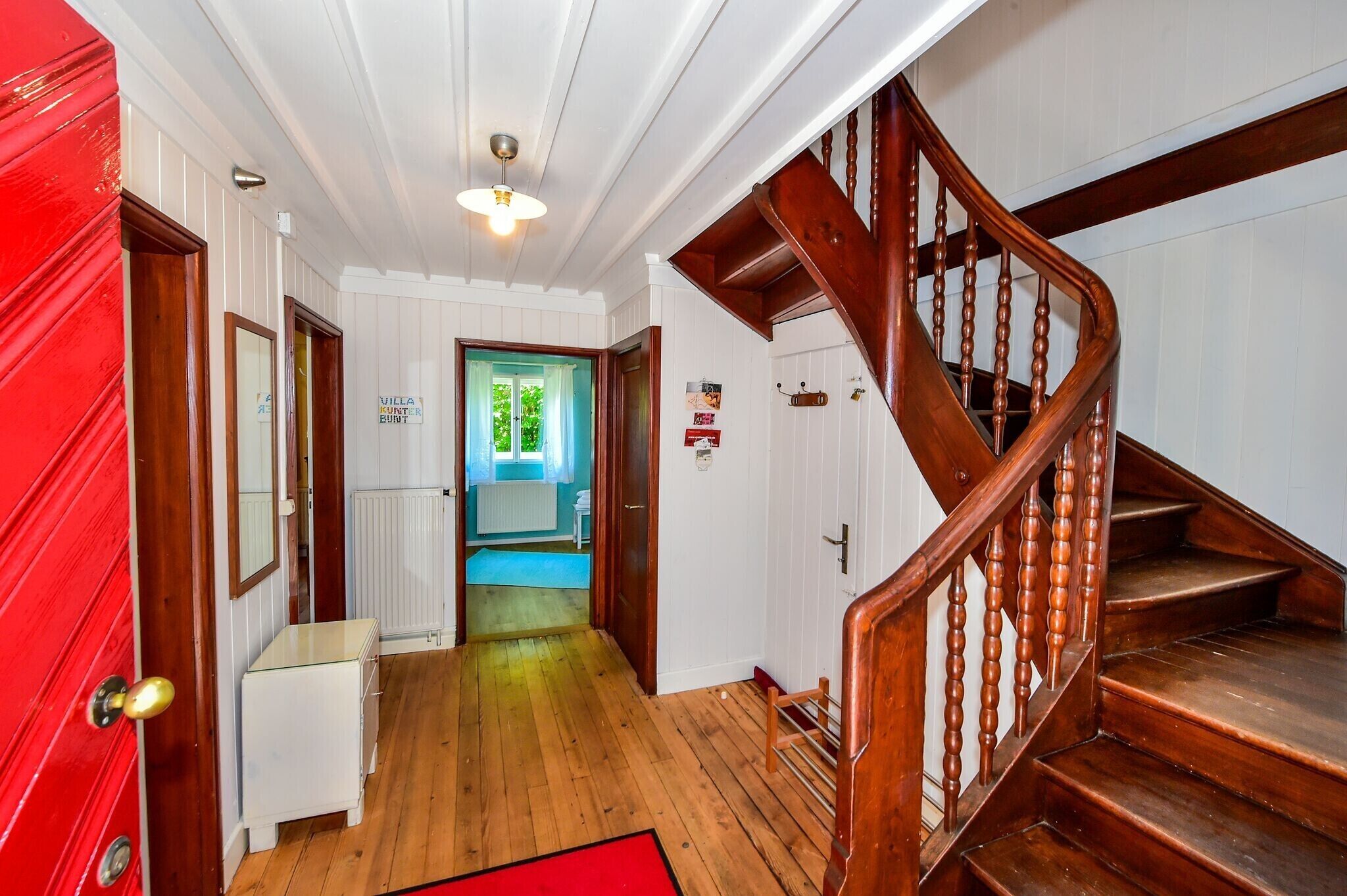 Una escalera de madera que lleva al segundo piso, un pasillo con paredes blancas y pisos de madera, y una puerta roja a la izquierda.