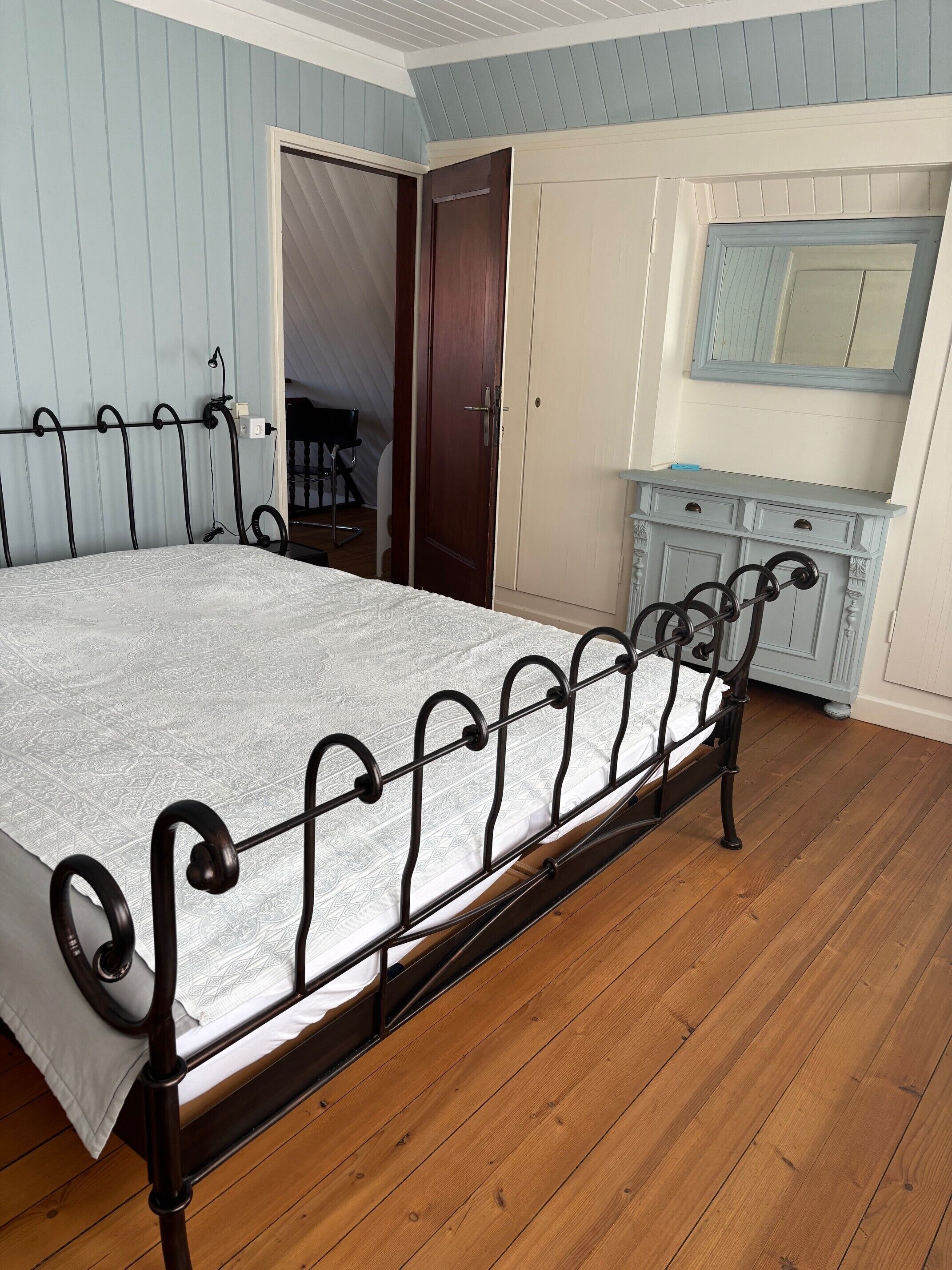 Een slaapkamer met een metalen bedframe, houten vloer, een kledingkast en een spiegel.