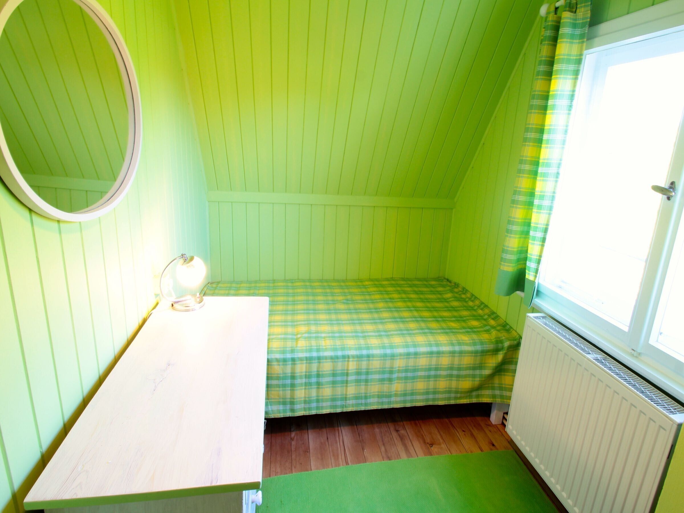 Una habitación pequeña con decoración verde, una cama individual, un espejo redondo, una mesita y una ventana con cortinas.
