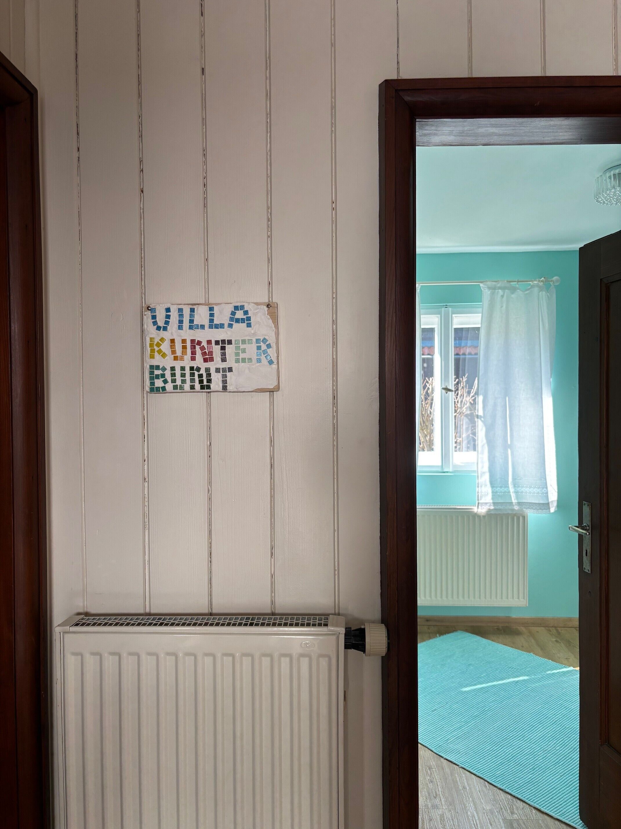 Ein Zimmer mit Heizkörper, einer Wandtafel, einem Fenster mit Vorhängen und einer Tür.