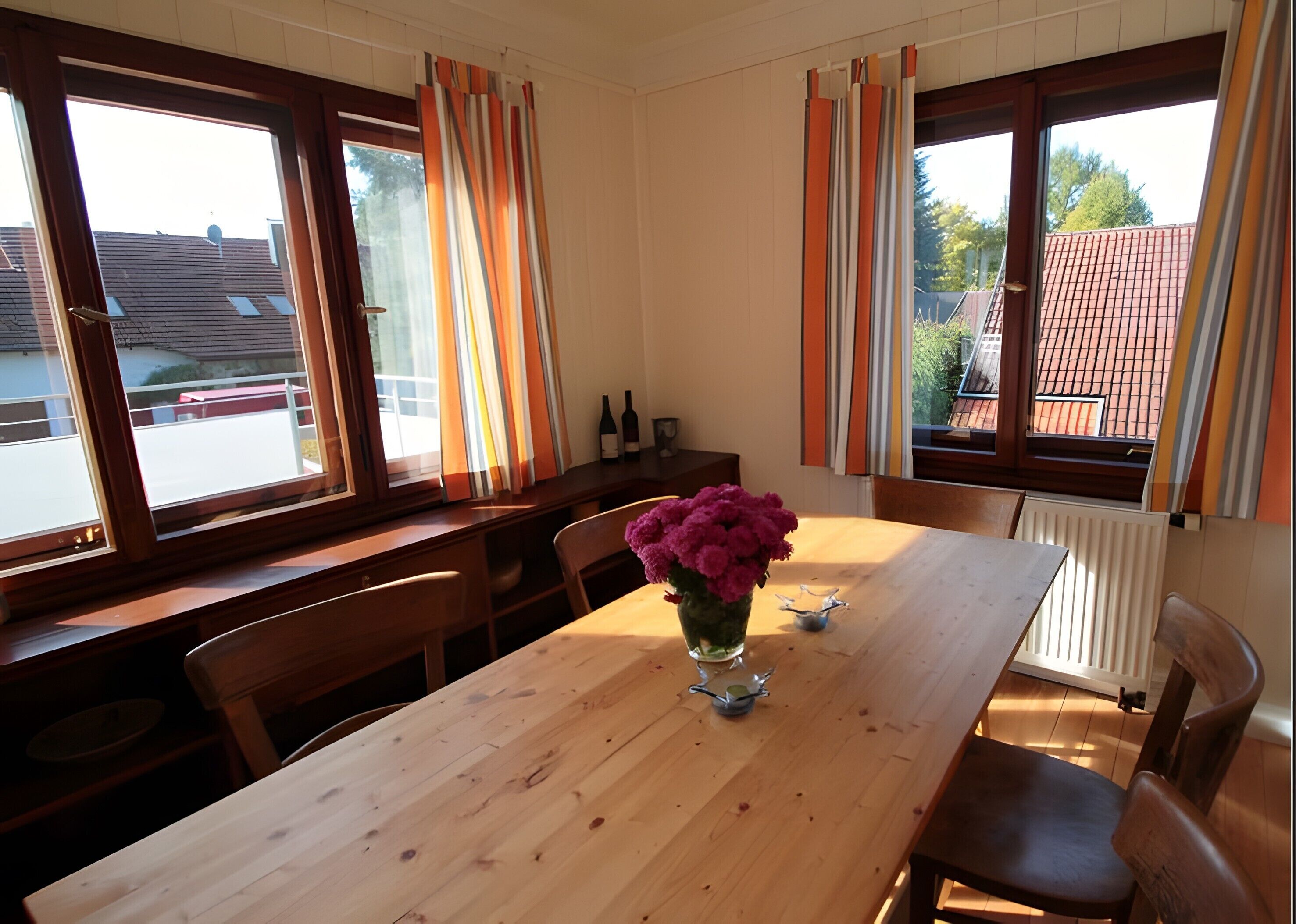 Un comedor con una mesa de madera, sillas, un florero con flores y ventanas con cortinas.