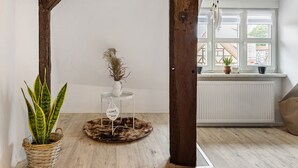 Een kamer met een plafond met houten balken, een witte radiator, een raam met uitzicht op gebouwen, een ronde tafel met een plant en een vaas, en een geweven mand met een plant.