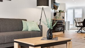 Een moderne woonkamer met een houten salontafel, een zwarte vaas met groene planten, een grijze bank met een groen kussen en een zwarte vloerlamp.