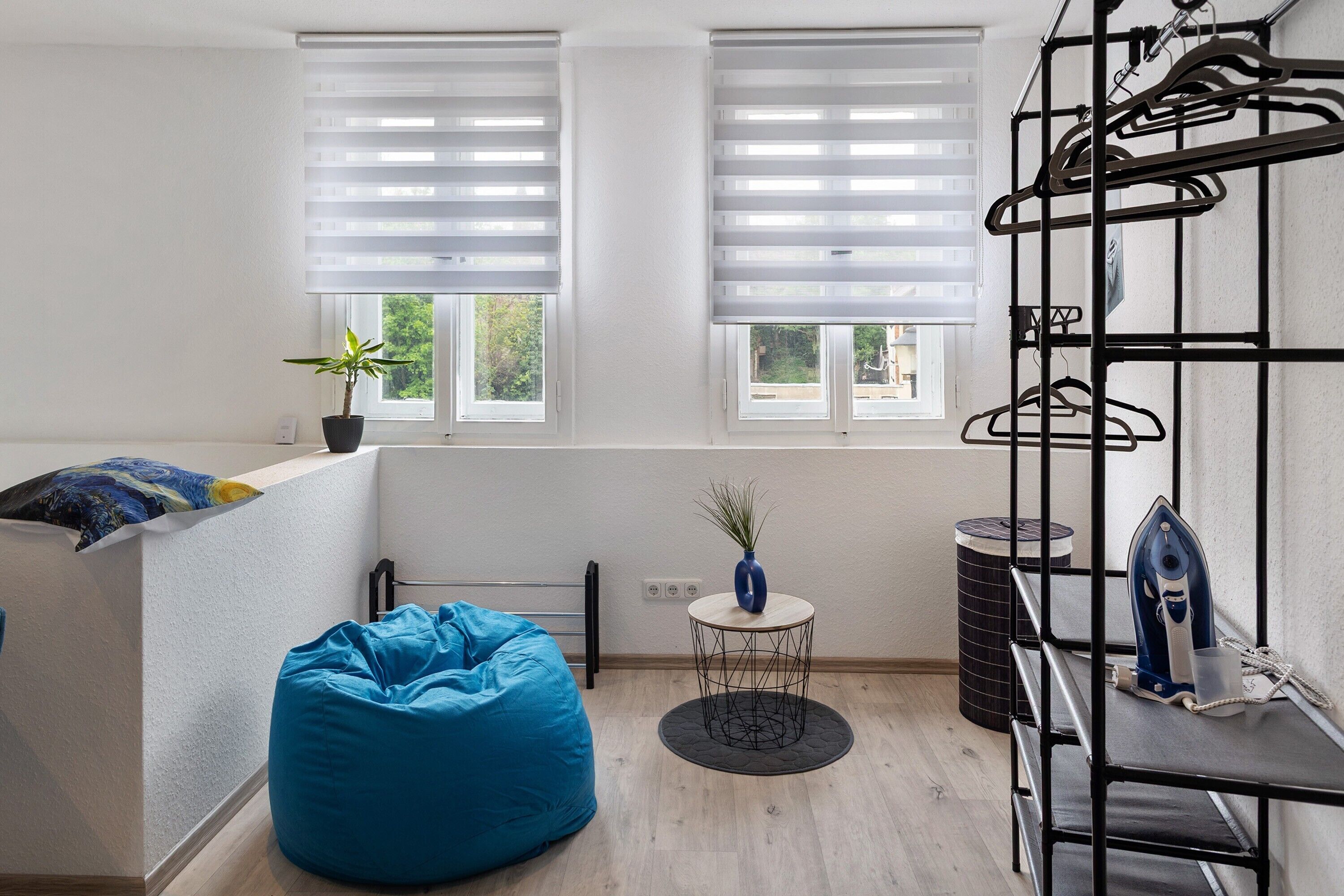 A room with a bed, a blue bean bag, a small table with a plant, and a metal rack with hangers.