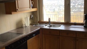 Une cuisine avec des meubles en bois, un lave-vaisselle, une grande fenêtre donnant sur un quartier résidentiel et un luminaire suspendu.