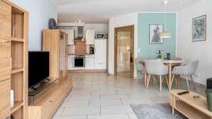 A modern living room with a TV, wooden shelves, a dining area with chairs, a kitchen in the background, and a light blue wall. - Apartment 'Schilfbrise' with Balcony and Wi-Fi (Wunstorf)