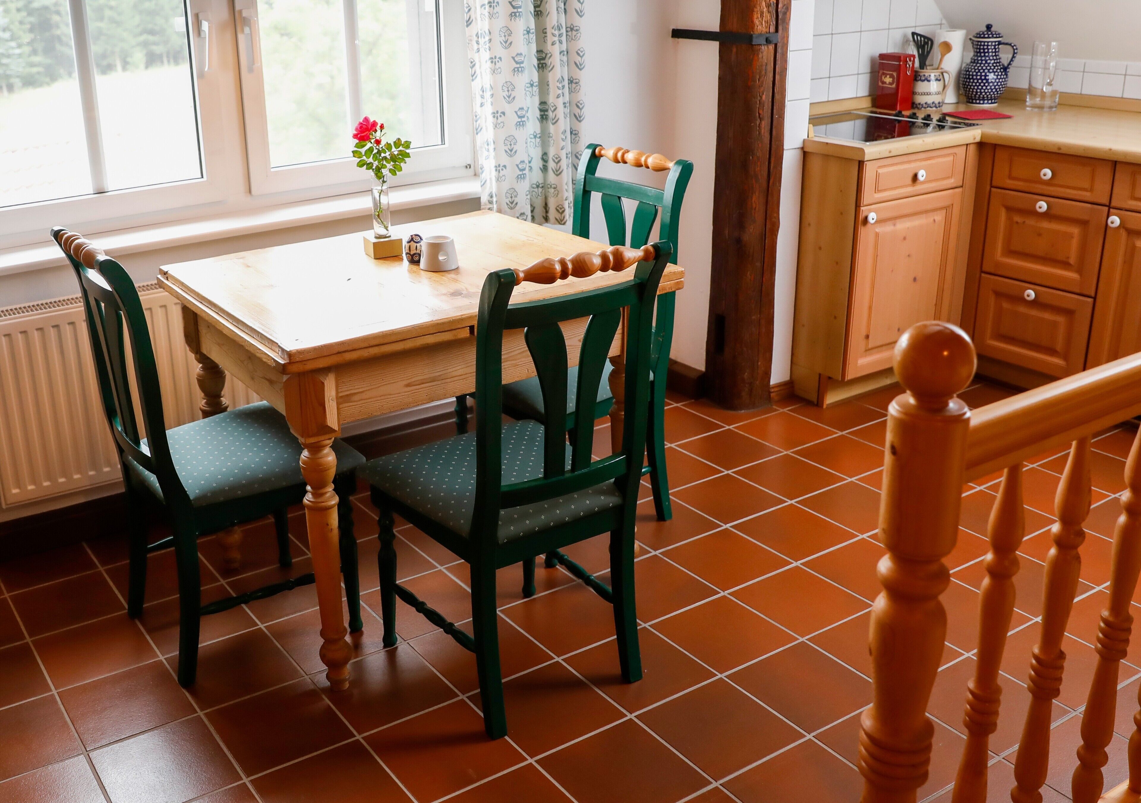 A kitchen with a wooden table, two green chairs, a window with curtains, and a tiled floor.