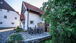 Une cour avec une terrasse en pierre, une table et des chaises, et une pile de bois de chauffage.