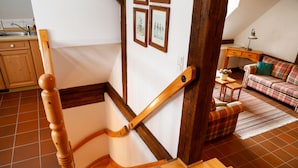Un escalier avec des balustrades en bois, un coin cuisine avec des placards en bois, un canapé à carreaux et une petite table avec une lampe.