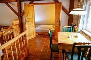 Una habitación con escalera de madera, cama con edredón blanco, mesa de comedor de madera con sillas y una ventana con cortinas.