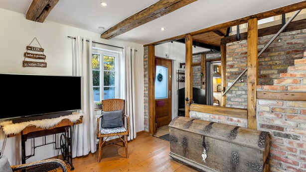 A room with a wooden ceiling, a television on a stand, a wicker chair, a wooden chest, and a brick wall.