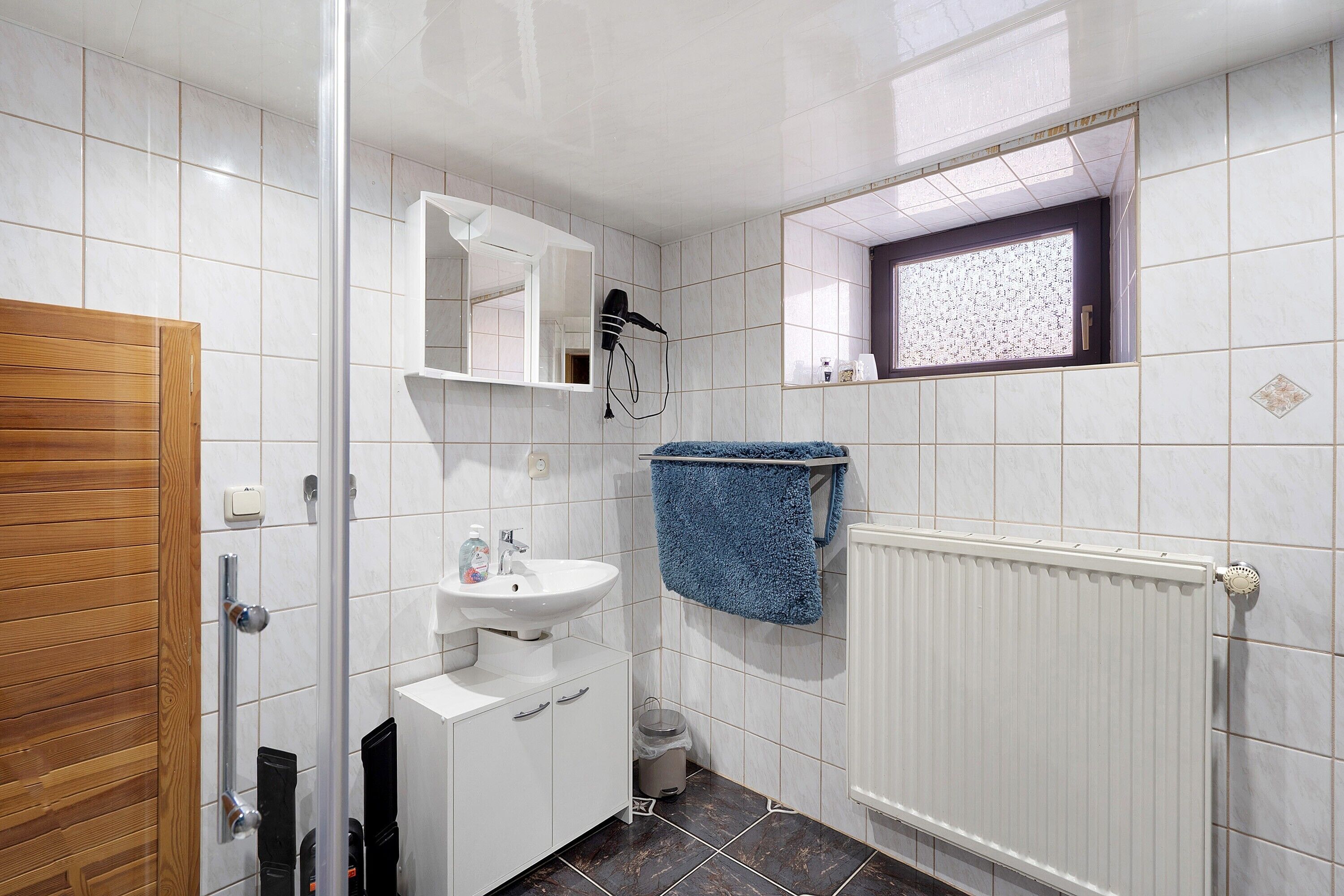 A bathroom with a wooden door, white tiles, a sink, a mirror, a towel rack, and a window.