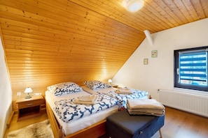 A bedroom with a sloped ceiling, wooden flooring, two beds with patterned bedding, a nightstand with a lamp, and a window with blinds.