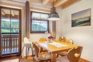 Un comedor con una mesa de madera, sillas, una lámpara colgante, una ventana con vistas a un bosque y un cuadro en la pared.