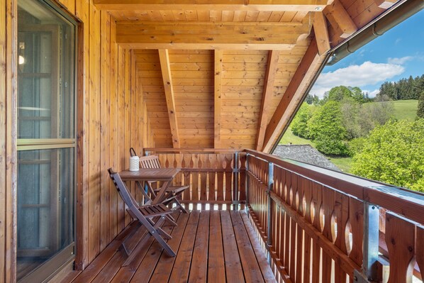 Un balcón de madera con una mesa y sillas, vistas a zonas verdes y un cielo despejado.