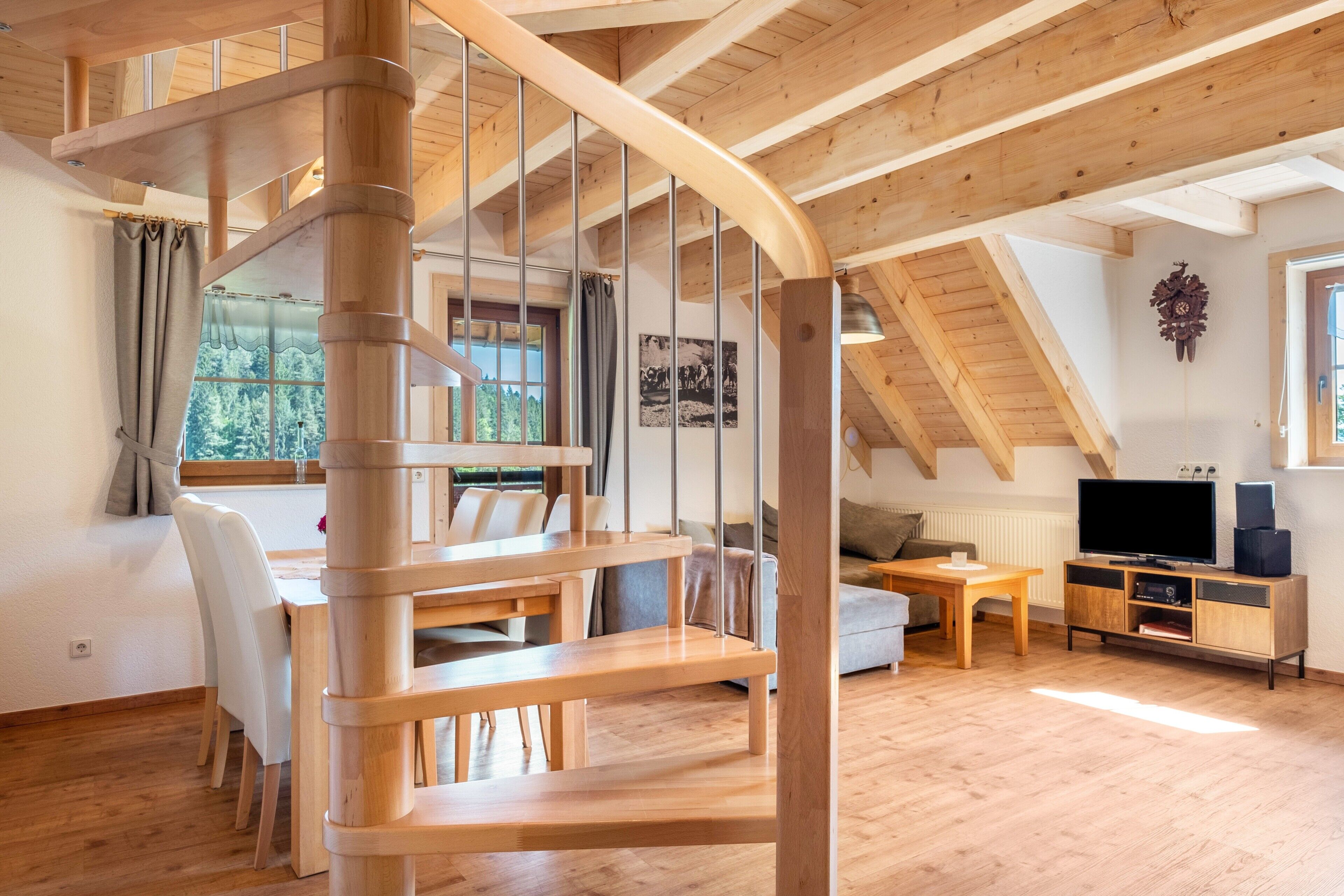 A wooden interior with a spiral staircase, a dining area, a living room with a TV, and a window with a view of trees.