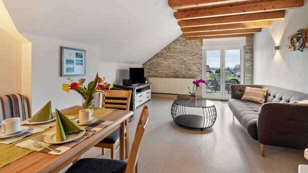 Un comedor con una mesa para dos personas, un sofá y un televisor en una habitación con techo inclinado y pared de piedra.