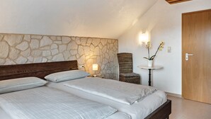Un dormitorio con pared de piedra, techo de madera, cama con ropa de cama blanca, una silla de mimbre, una mesita con una planta y una puerta de madera.