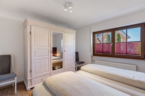 A bedroom with two beds, a wardrobe, a chair, and a window with a view of a house.
