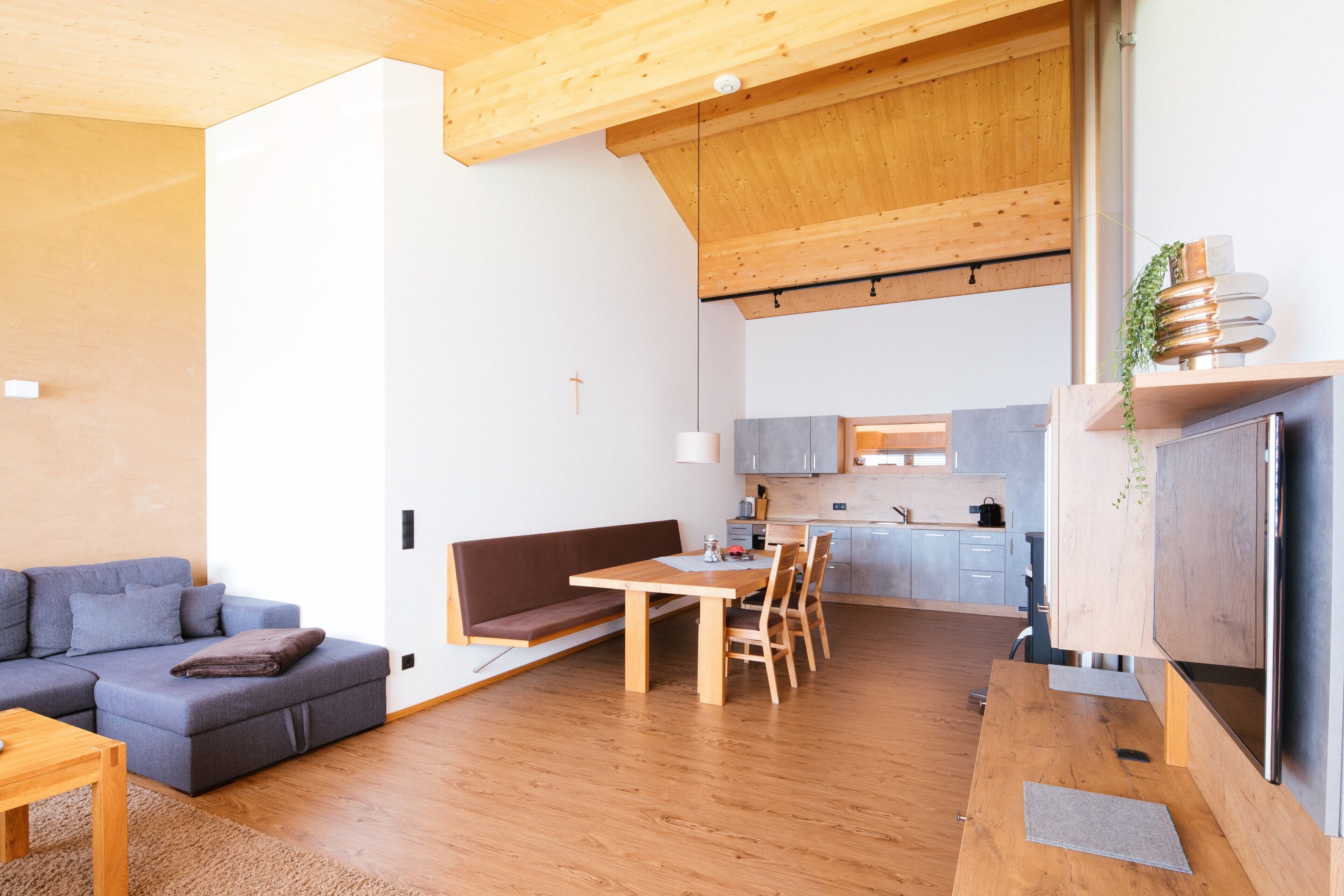 Et moderne interiør med træloft, spiseområde med bord og stole, en sofa og et køkken i baggrunden.