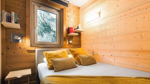 Une pièce de style cabane en bois, avec un lit, une fenêtre donnant sur une forêt, et des étagères remplies de livres et une lampe.