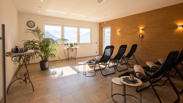 Een zonnige kamer met een houten wand, een groot raam, een klok en meerdere fauteuils.