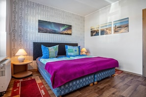 A bedroom with a bed, two bedside lamps, a nightstand, a red rug, patterned wallpaper, and a large window.