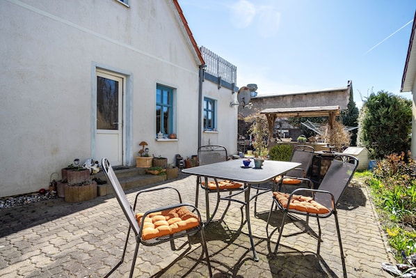 An outdoor patio with a table and chairs, a small garden area, and a building with a white exterior and blue windows.
