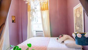 Un dormitorio con cama, ventana con cortinas, candelabro y un cuadro en la pared.