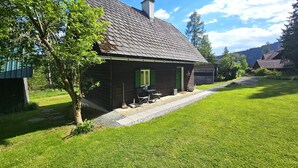 Una casa de madera rústica con techo inclinado, una chimenea y un pequeño porche con mobiliario exterior.