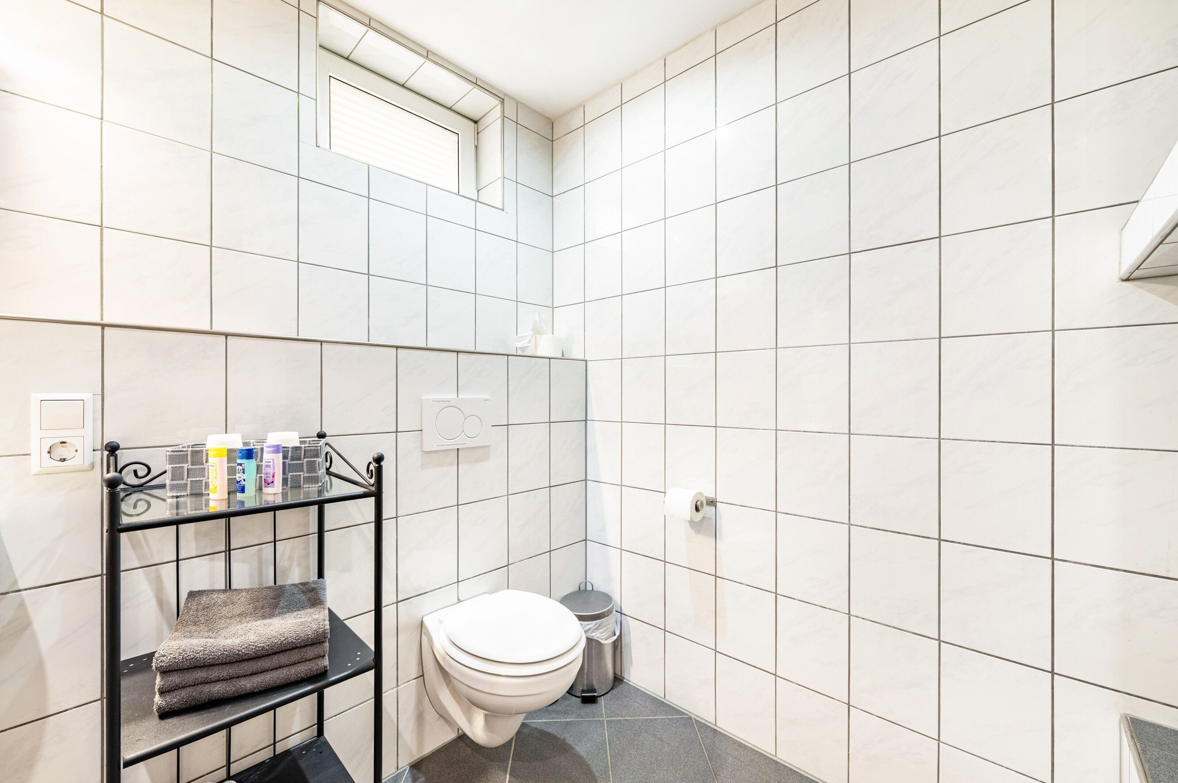 Un baño con paredes de azulejos blancos, un inodoro, un estante con toallas dobladas y un cubo de basura.