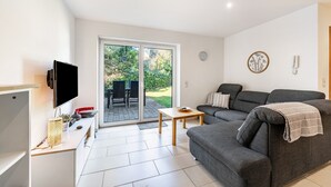 A modern living room with a flat-screen TV, a grey sofa, a wooden coffee table, and a sliding glass door leading to an outdoor patio. - Holiday Apartment 'Eifel im Herzen' with Private Terrace, Garden & Wi-Fi (Hambuch)