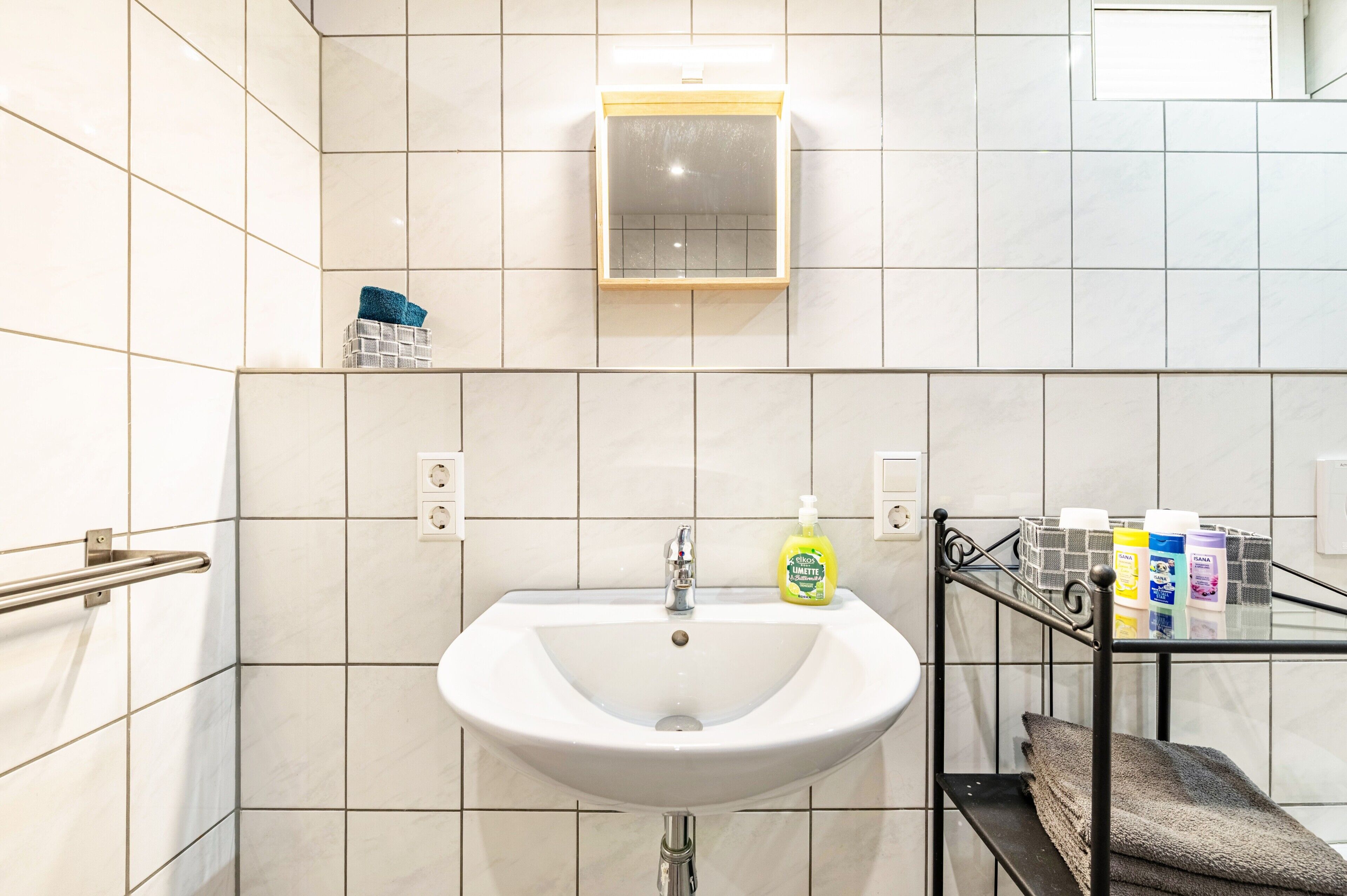 Kylpyhuone, jossa on valkoinen pesuallas, peili ja lasihylly, jossa on hygieniatuotteita.