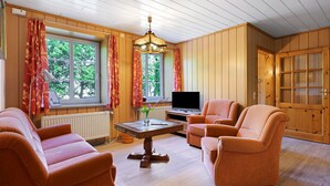 A cozy living room with wooden walls, a large window with curtains, a television, and two orange sofas. - Apartment 'Norderoog' with Private Terrace, Shared Garden and Wi-Fi (Nordstrand)