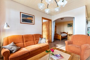 A living room with a sofa, armchair, and a coffee table with a vase. There is a wooden door leading to another room.