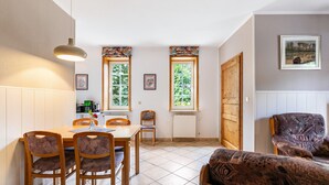 A dining area with a wooden table and chairs, two windows with curtains, a radiator, and a sofa. - Apartment 'Hooge' with Private Terrace, Shared Garden and Wi-Fi (Nordstrand)
