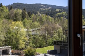 Une vue depuis une fenêtre donnant sur une colline boisée, un bâtiment avec un toit en pente et une porte dotée d’une poignée.