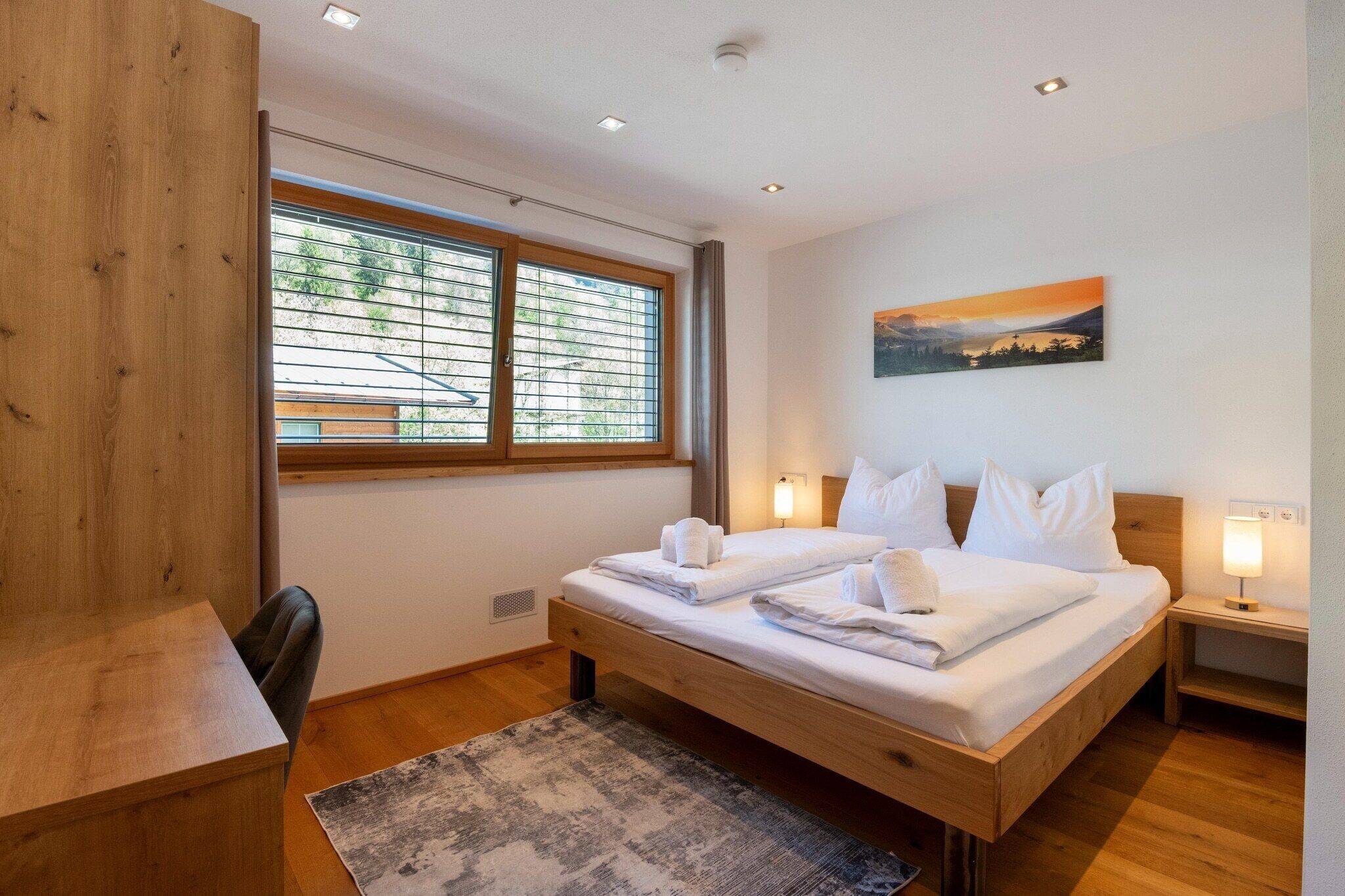 Une chambre d’hôtel avec un lit en bois, un bureau, une chaise, une fenêtre avec des stores, un tableau au mur et un tapis sur le sol.