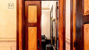 A wooden door with a sign reading "3-C APARTMENT" leading to a hallway with a chair and a trash bin.