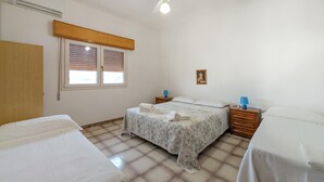 Ein Schlafzimmer mit zwei Betten, einem Deckenventilator, einem Fenster mit Jalousien, einem Nachttisch mit Lampe und einem an der Wand hängenden Bild.