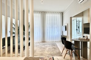 Una sala de estar moderna con un sofá, una mesa de centro y un televisor. Cuenta con una zona de comedor con una mesa y sillas. La habitación tiene ventanas amplias con cortinas ligeras.