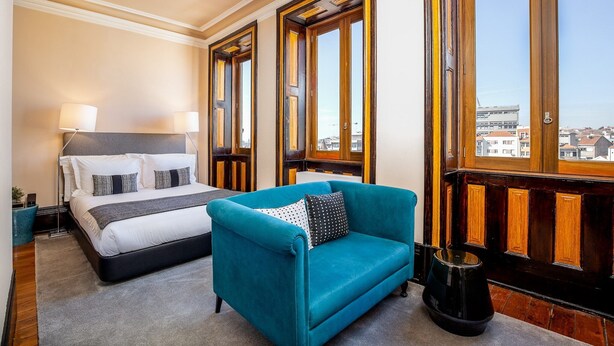 A hotel room with a bed, a blue armchair, and large windows with wooden frames.