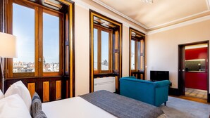A hotel room with a bed, a blue armchair, a TV, and large windows offering a city view.