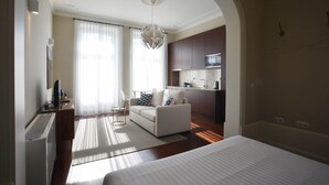 A modern hotel room with a large bed, a sitting area with a sofa and a small table, a kitchenette, and a large window with sheer curtains.