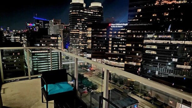 Un balcón con dos sillas y una mesa, con vistas a un paisaje urbano nocturno con edificios iluminados y una vista despejada de las calles de abajo.
