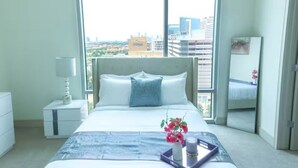 Habitación de hotel con una cama grande, una mesita de noche con lámpara, una mesita auxiliar con un jarrón de flores y vistas de la ciudad a través de amplios ventanales.