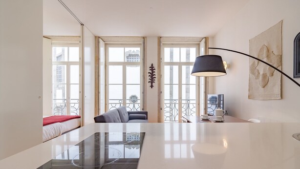 A modern living room with a glass dining table, a sofa, and a coffee table. There are large windows with a view of buildings outside.