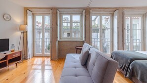 Una habitación moderna con un sofá gris, un televisor sobre un mueble de madera y una ventana con vistas a edificios.