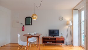 Una sala de estar moderna con una mesa de comedor de madera, sillas blancas, un televisor sobre un mueble de madera, una lámpara colgante y una lámpara de pie.